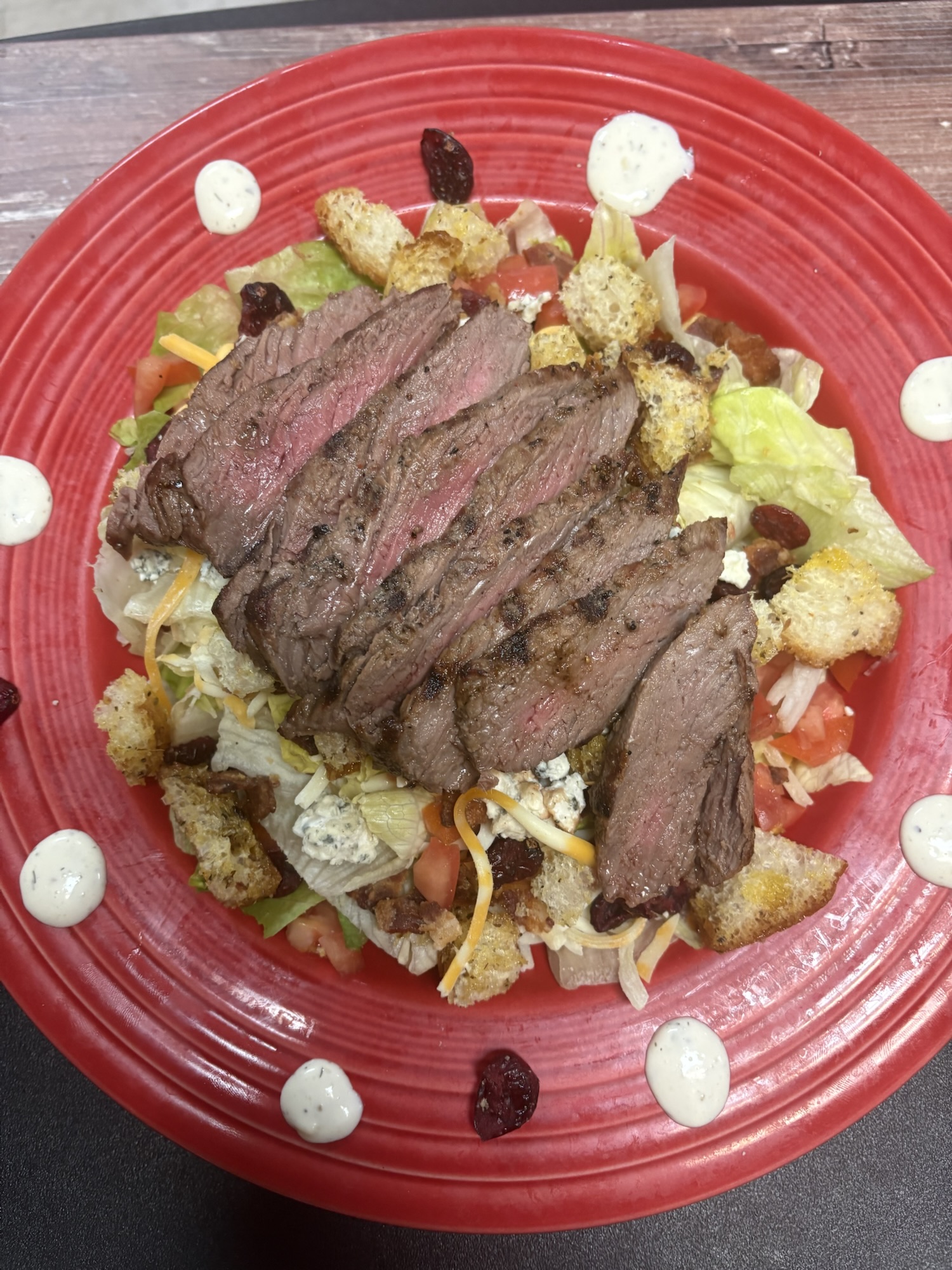 A vibrant red plate featuring a salad topped with sliced grilled steak, croutons, shredded cheese, tomatoes, and lettuce, garnished with dollops of dressing.